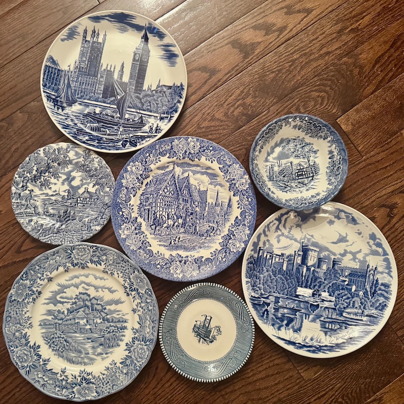 May include: A collection of six ceramic plates and one bowl featuring blue and white transferware patterns. The designs include detailed illustrations of Big Ben, Windsor Castle, rural landscapes, and historic buildings with intricate floral borders.