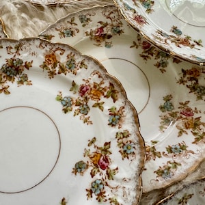 K&ouml;nnte beinhalten: Ein Set vintage Porzellan Teller mit floralen Mustern und goldenen Verzierungen. Die Teller haben einen gewellten Rand und sind &uuml;bereinander gestapelt.