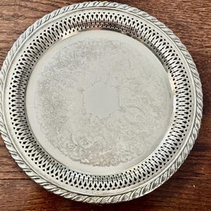 May include: A round silver-toned serving tray featuring a decorative openwork lattice border with a twisted rope-style edge. The center of the tray displays an intricate, etched floral pattern surrounding a smooth, plain hexagonal area.
