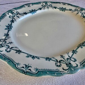 May include: An oval ceramic platter featuring a cream-coloured centre and a scalloped edge. The rim is decorated with a dark teal transferware pattern consisting of floral garlands, intricate ribbons, and symmetrical scrollwork motifs.
