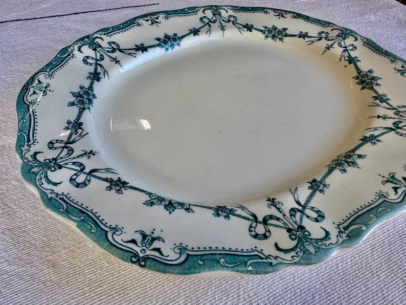 May include: An oval ceramic platter featuring a cream-coloured centre and a scalloped edge. The rim is decorated with a dark teal transferware pattern consisting of floral garlands, intricate ribbons, and symmetrical scrollwork motifs.