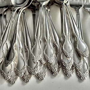 May include: Close-up of a collection of ornate silver silverware, including forks and spoons. The silverware has a floral design on the handles and a shiny, reflective surface. The arrangement suggests a formal dining setting.