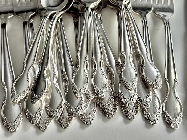 May include: Close-up of a collection of ornate silver silverware, including forks and spoons. The silverware has a floral design on the handles and a shiny, reflective surface. The arrangement suggests a formal dining setting.
