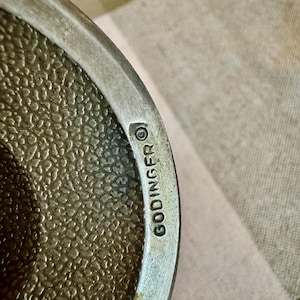 May include: A metallic object with a textured, pebbled surface and a smooth, polished rim. The word GODINGER is stamped into the metal along the edge, accompanied by a copyright symbol. The finish appears to be silver-toned with a matte appearance.