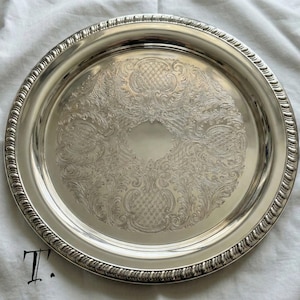 May include: A round silver-toned serving tray featuring an intricate engraved pattern of scrolling leaves and cross-hatched details in the center. The tray has a raised, decorative rope-style border along the outer edge and a smooth, polished finish.