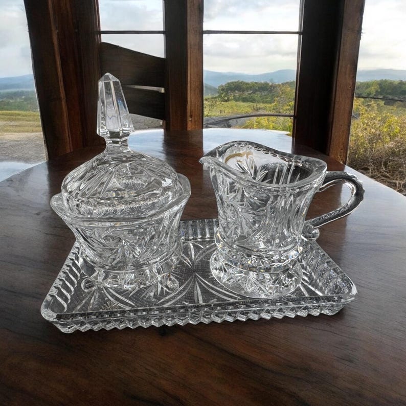 May include: A clear glass creamer and a lidded sugar bowl set with intricate starburst and geometric patterns, both resting on a matching rectangular glass tray. The pieces feature faceted edges and a brilliant, reflective finish throughout.