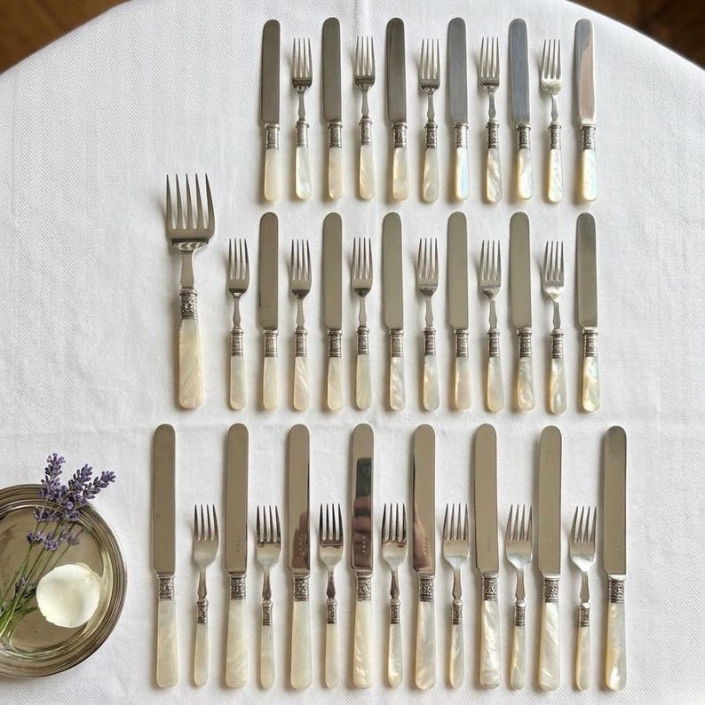 May include: An assortment of antique silverware, including forks and knives, arranged on a white tablecloth. The silverware features ornate silver handles with a pearlescent finish, creating an elegant and vintage aesthetic. A small silver tray with lavender and a petal is in the lower left corner.