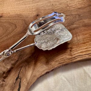 May include: Silver serving tongs with an ornate design on the handle and a decorative, engraved, silver serving spoon attached to the tongs.