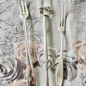May include: Three silver serving utensils on a floral patterned cloth. The utensils include two forks and a silver cake server with a handle and a clamp. The forks have three tines each. The cloth has a pattern of roses in shades of pink and beige.