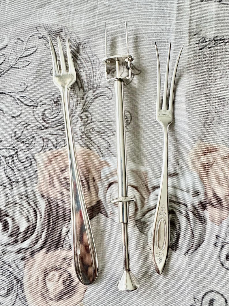 May include: Three silver serving utensils on a floral patterned cloth. The utensils include two forks and a silver cake server with a handle and a clamp. The forks have three tines each. The cloth has a pattern of roses in shades of pink and beige.
