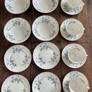 May include: A collection of white porcelain dishware with a floral pattern in blue and green. The set includes bowls, plates, and teacups with saucers, all featuring a gold trim. The arrangement is on a wooden surface.