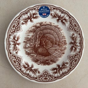 Op de afbeelding: Een rond wit keramieken bord met een bruine transferprint van een kalkoen in een landelijk landschap. De rand is versierd met een herhalend patroon van eikenbladeren en eikels. Een blauwe sticker vermeldt Royal Stafford Est 1845 Made in England.