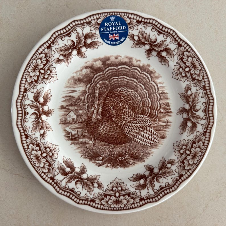 Op de afbeelding: Een rond wit keramieken bord met een bruine transferprint van een kalkoen in een landelijk landschap. De rand is versierd met een herhalend patroon van eikenbladeren en eikels. Een blauwe sticker vermeldt Royal Stafford Est 1845 Made in England.