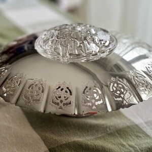 May include: A silver-colored serving dish with a decorative, scalloped edge and intricate cut-out designs. A clear, glass lid with a detailed relief pattern rests on top. The dish is on a patterned surface.