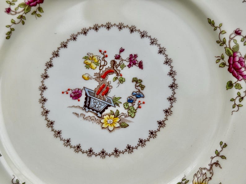 Puede incluir: Un plato de cer&aacute;mica blanco con un dise&ntilde;o floral. El centro del plato presenta un motivo floral estilizado con una planta en maceta en el centro. El plato tiene un borde festoneado marr&oacute;n.