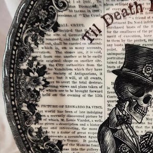 K&ouml;nnte beinhalten: Ein schwarzer und wei&szlig;er Keramikteller mit einem dekorativen Rand und einem Sch&auml;del, der einen Zylinder und einen Anzug tr&auml;gt. Der Teller enth&auml;lt Text, der "Til Death" und "ALL CRYPT" lautet.