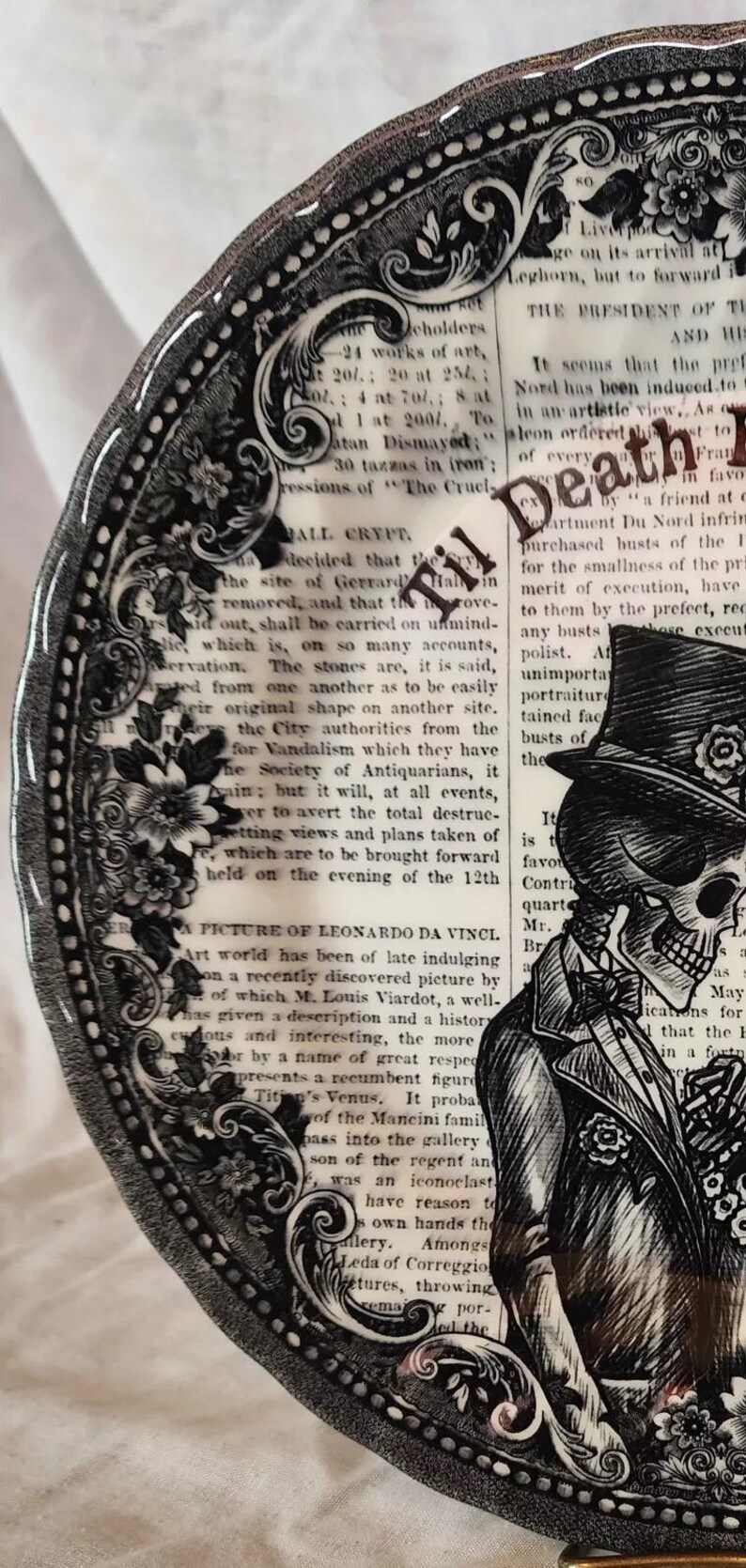 K&ouml;nnte beinhalten: Ein schwarzer und wei&szlig;er Keramikteller mit einem dekorativen Rand und einem Sch&auml;del, der einen Zylinder und einen Anzug tr&auml;gt. Der Teller enth&auml;lt Text, der "Til Death" und "ALL CRYPT" lautet.