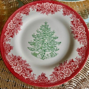 May include: A white ceramic plate with a red floral border and a green Christmas tree design in the center. The tree is decorated with green ornaments and sits above wrapped gifts. The plate is a festive holiday item.