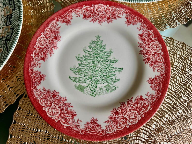 May include: A white ceramic plate with a red floral border and a green Christmas tree design in the center. The tree is decorated with green ornaments and sits above wrapped gifts. The plate is a festive holiday item.