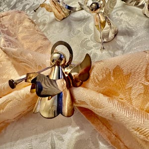 K&ouml;nnte beinhalten: Silberne Serviettenringe in Form von Engeln mit Trompetendetails, auf einer pfirsichfarbenen Serviette. Die Engelsfiguren haben detaillierte Fl&uuml;gel und Heiligenscheine. Der Hintergrund zeigt eine wei&szlig;e Tischdecke mit Muster, die eine festliche und elegante Tischdekoration schafft.