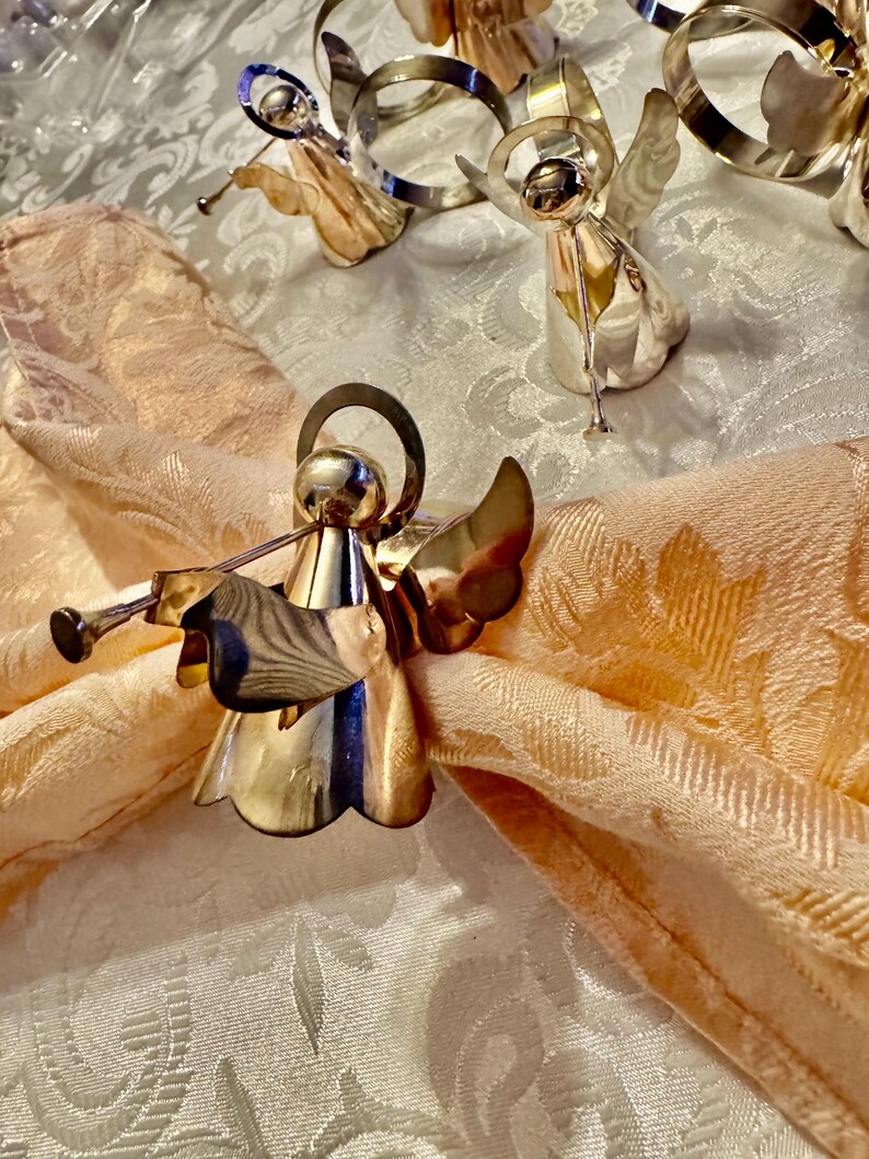 K&ouml;nnte beinhalten: Silberne Serviettenringe in Form von Engeln mit Trompetendetails, auf einer pfirsichfarbenen Serviette. Die Engelsfiguren haben detaillierte Fl&uuml;gel und Heiligenscheine. Der Hintergrund zeigt eine wei&szlig;e Tischdecke mit Muster, die eine festliche und elegante Tischdekoration schafft.