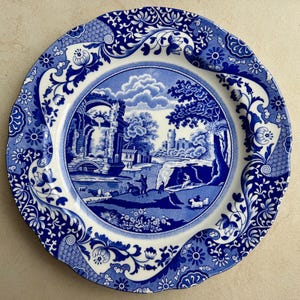 Puede incluir: Un plato redondo de cer&aacute;mica con un patr&oacute;n azul y blanco. El centro representa un paisaje pastoral con ruinas, &aacute;rboles y figuras, mientras que el borde festoneado est&aacute; decorado con intrincados motivos florales y de volutas.