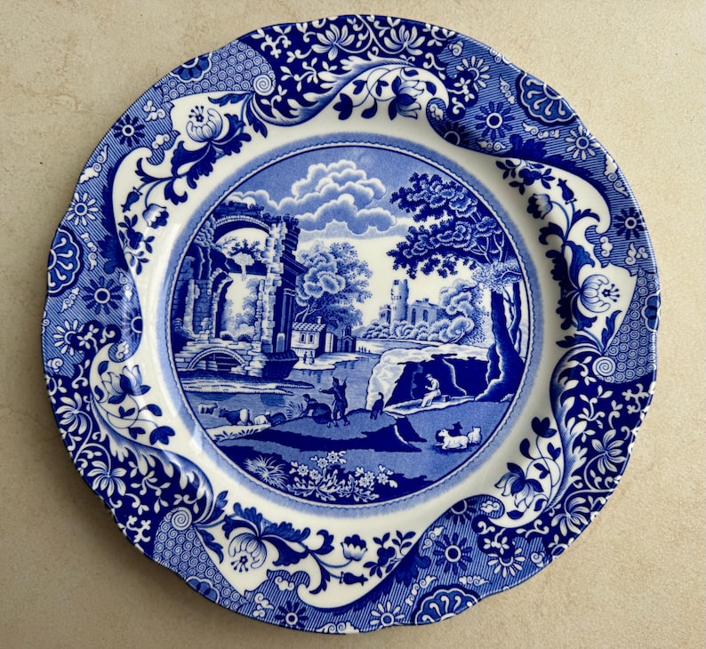 Puede incluir: Un plato redondo de cer&aacute;mica con un patr&oacute;n azul y blanco. El centro representa un paisaje pastoral con ruinas, &aacute;rboles y figuras, mientras que el borde festoneado est&aacute; decorado con intrincados motivos florales y de volutas.
