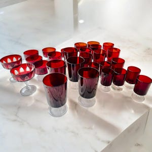 May include: A collection of 20 red glass goblets and a red glass coupe. The goblets are all the same size and shape, and the coupe is smaller. The goblets have a clear glass base and a red glass bowl. The coupe has a clear glass stem and a red glass bowl.