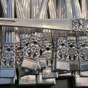 May include: A collection of silver-toned metal flatware handles arranged in a parallel stack. Each handle features a vertical ribbed design and a decorative floral relief pattern at the base. One handle is stamped with the word COMMUNITY.