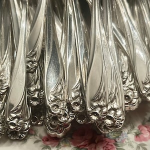 Peut inclure: Gros plan sur une collection de couverts argentés, probablement des fourchettes, avec un motif floral à la base des manches. Les couverts sont disposés de près, reflétant la lumière. Le motif floral ajoute un élément décoratif aux ustensiles.