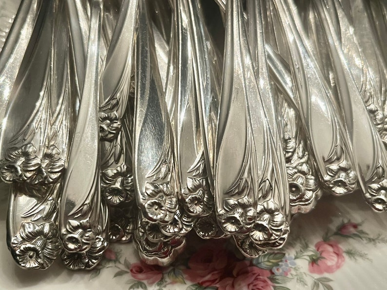 May include: A collection of silver-toned metal spoons featuring a raised floral daffodil motif on the handles. The handles have a smooth, tapered design with detailed embossed flower patterns at the base of each individual spoon handle.
