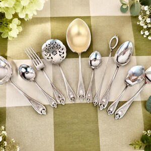 May include: A collection of silver-toned flatware including a large serving spoon, a slotted spoon with floral cutouts, a fork, several spoons of varying sizes, and a long-handled stirrer, all featuring ornate embossed detailing on the handles.