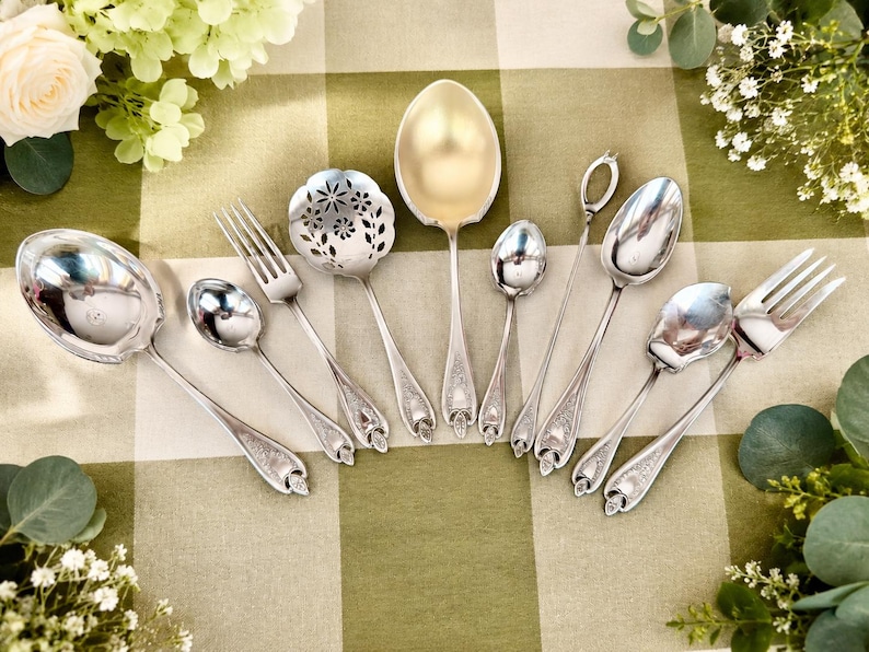 May include: A collection of silver-toned flatware including a large serving spoon, a slotted spoon with floral cutouts, a fork, several spoons of varying sizes, and a long-handled stirrer, all featuring ornate embossed detailing on the handles.