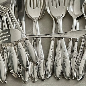 May include: A collection of silver-colored flatware, including forks and spoons, with a floral design on the handles. One fork is in focus, displaying the word "Springtime". The flatware is arranged on a light-colored surface.