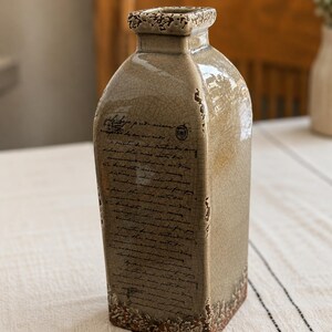 May include: A rectangular, ceramic vase with a distressed, crackled finish in shades of tan and brown. The vase features a square opening and is adorned with faded script. The base and rim have a textured, aged appearance, adding to its vintage aesthetic.