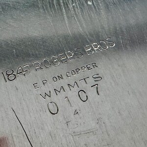 Może przedstawiać: Srebrzysta metalowa powierzchnia z wygrawerowanym znakiem producenta o treści 1847 ROGERS BROS, E.P. ON COPPER, WMMTS oraz 0107. Metalowe wykończenie wykazuje drobne liniowe zarysowania oraz odblaskową, polerowaną teksturę na płaskim obszarze.