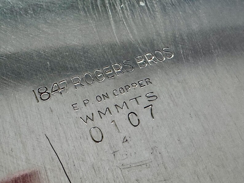 Może przedstawiać: Srebrzysta metalowa powierzchnia z wygrawerowanym znakiem producenta o treści 1847 ROGERS BROS, E.P. ON COPPER, WMMTS oraz 0107. Metalowe wykończenie wykazuje drobne liniowe zarysowania oraz odblaskową, polerowaną teksturę na płaskim obszarze.