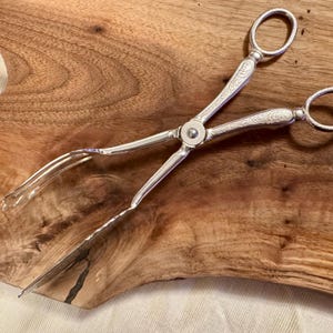 May include: Silver-plated serving tongs with an ornate design. The tongs have a long, slender handle and a pair of prongs at the end.