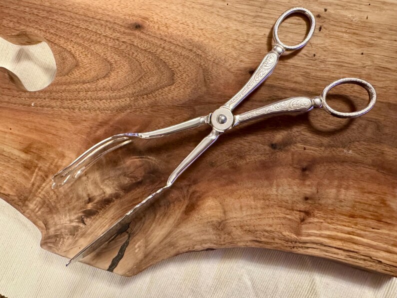 May include: Silver-plated serving tongs with an ornate design. The tongs have a long, slender handle and a pair of prongs at the end.