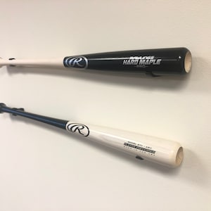 May include: Two baseball bats mounted on a white wall. The top bat is black with white lettering that reads "Rawlings Hard Maple - Pro". The bottom bat is white with black lettering that reads "Model 571 - Ash Player Preferred Made in the USA".