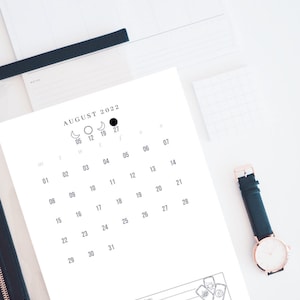 May include: A black and white calendar for August 2022 with moon phases and a space for monthly manifestation. The calendar has a simple design with a white background and black text.