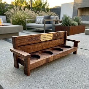 Op de afbeelding: Een houten drankhouder in de vorm van een bank met vier cirkelvormige uitsparingen. Het donkerbruine hout heeft een decoratief ontwerp en een kleine gouden plaat met tekst. De bank heeft armleuningen en een rugleuning. De tekst op de plaat luidt: "Pop's Bench - Where stories are poured and songs are played Love Addison and Emma."