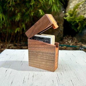 Peut inclure: Une boîte à cartes à jouer rectangulaire en bois de noyer brun foncé avec un accent en bois plus clair et texturé sur le côté et le bord supérieur. Le couvercle est articulé en angle, révélant un jeu de cartes noir et blanc à l'intérieur.