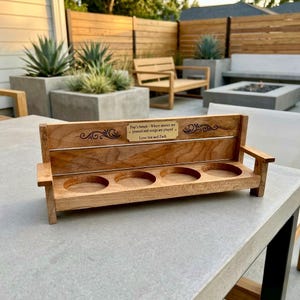 May include: A wooden bench-shaped flight paddle with four circular cutouts for glasses. A metal plaque on the backrest features the engraved text: Pop's bench - Where stories are poured and songs are played. Love Sue and Zach.