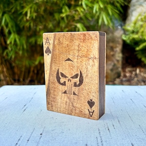 Peut inclure: Un étui rectangulaire en bois au grain brun clair, présentant un motif gravé au laser représentant un as de pique. Le centre affiche un emblème de crâne stylisé, avec la lettre A et un symbole de pique dans les coins supérieur gauche et inférieur droit.