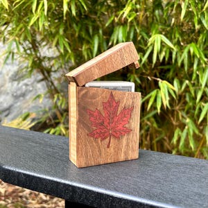 May include: A rectangular wooden playing card box with a hinged lid, crafted from light brown wood with a visible grain. A prominent, detailed red maple leaf graphic is centered on the front panel of the box, which holds a deck of cards.