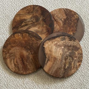 May include: A set of four circular wooden coasters. Each coaster displays a unique wood grain pattern in shades of brown and tan. The coasters have a dark brown border, providing contrast to the natural wood design. These coasters are designed to protect surfaces from drink marks.