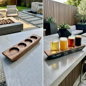 May include: A rectangular wooden beer flight paddle featuring four circular cutouts for glasses. The front edge is painted black with white chalk text reading Pilsner, Hazy IPA, Amber Ale, and Stout, holding four glasses of varying beer colors.