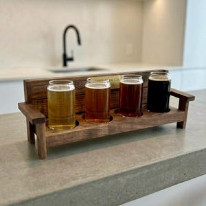 May include: A wooden beer flight set shaped like a bench, holding four glasses of beer. The beers vary in color from light gold to dark brown. The bench has a small engraved plaque that reads "Bench Brew Co. 26".