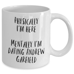 May include: White ceramic mug with the text "PHYSICALLY I'M HERE" above "MENTALLY I'M DATING ANDREW GARFIELD" in black, bold font. The mug has a curved handle and a smooth, glossy finish.